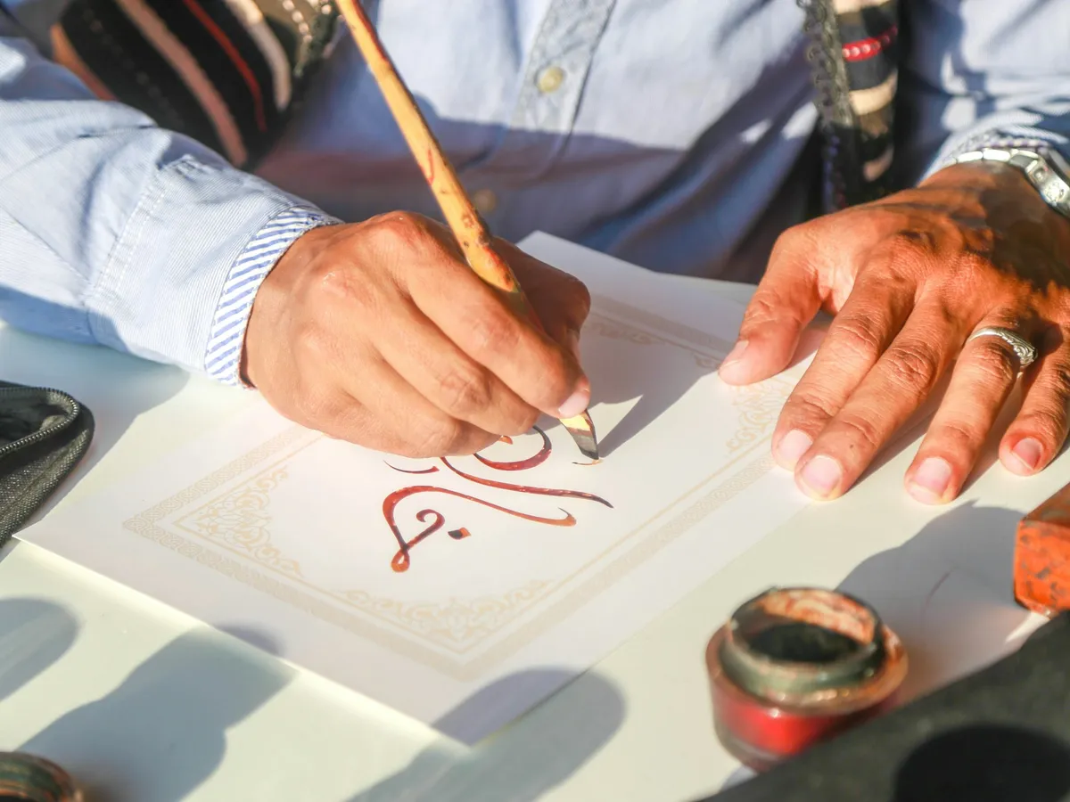 A hand forming Arabic letters with a reed pen, an inkwell beside the page.