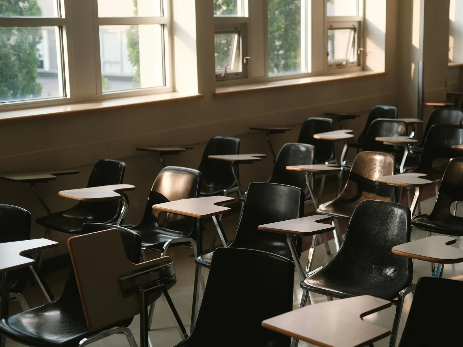 Rows of empty desks in a classroom, lit by a wall of tall windows.