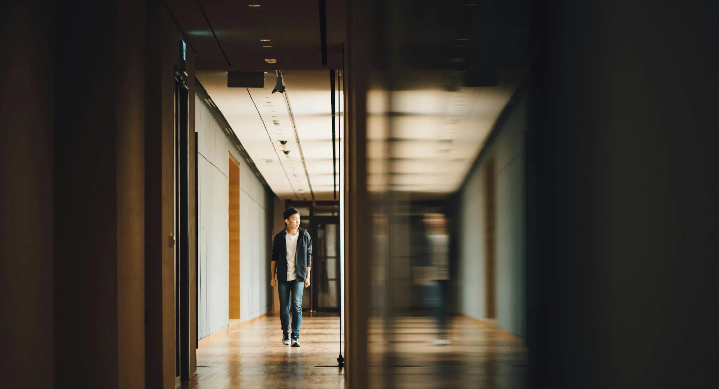 A school corridor with daylight falling across the floor and one person walking at the far end.