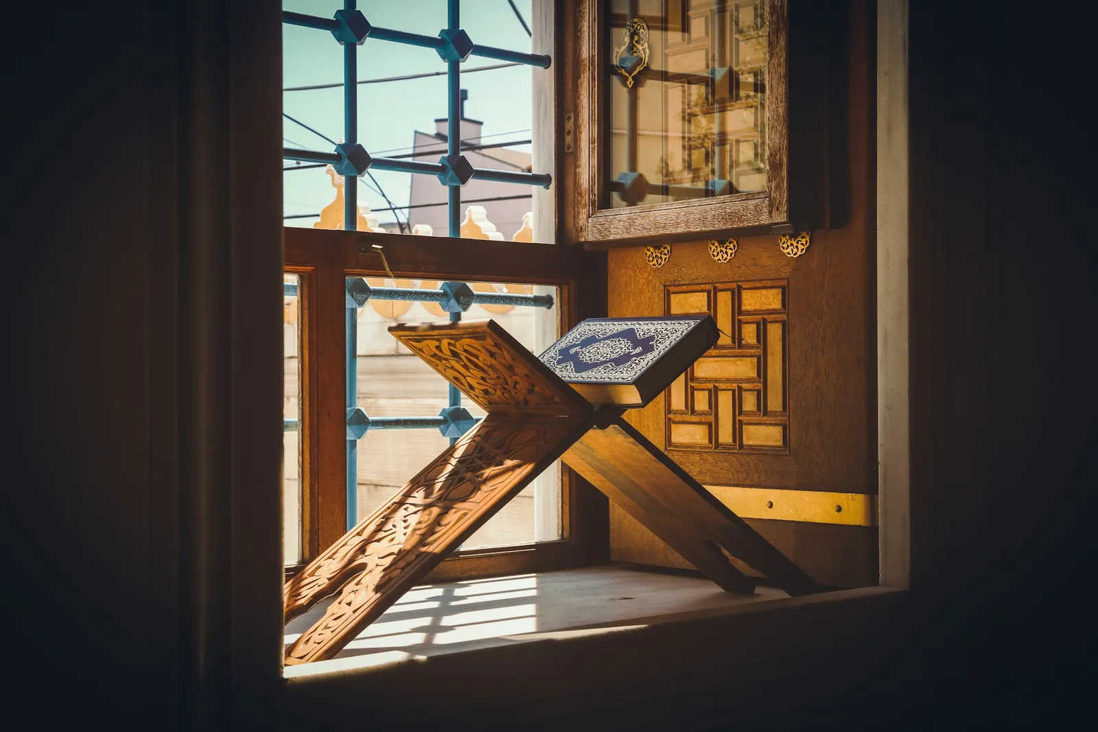 A mushaf open on a carved wooden rihal, with morning light coming through the window behind it.